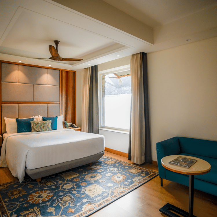 A brightly lit hotel room features a neatly made bed with white linens, a teal armchair, and a small round side table with a lamp. A window with sheer curtains allows natural light to filter in - Ananta Spa and Resort, Pushkar