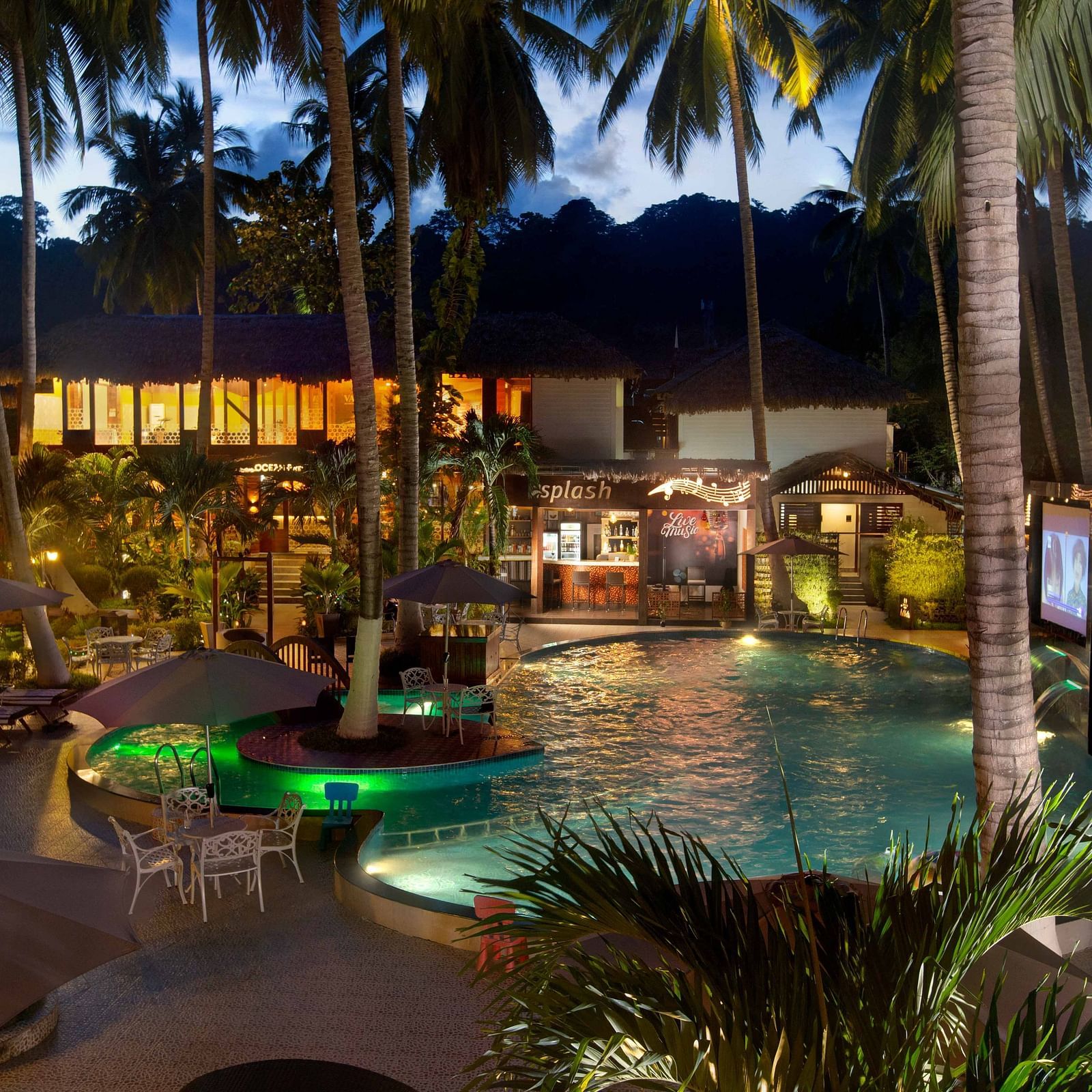  A night-time view of a resort pool area, illuminated by soft lighting. Palm trees surround the pool, creating a tropical and inviting atmosphere - Coral Reef Resort & Spa, Havelock