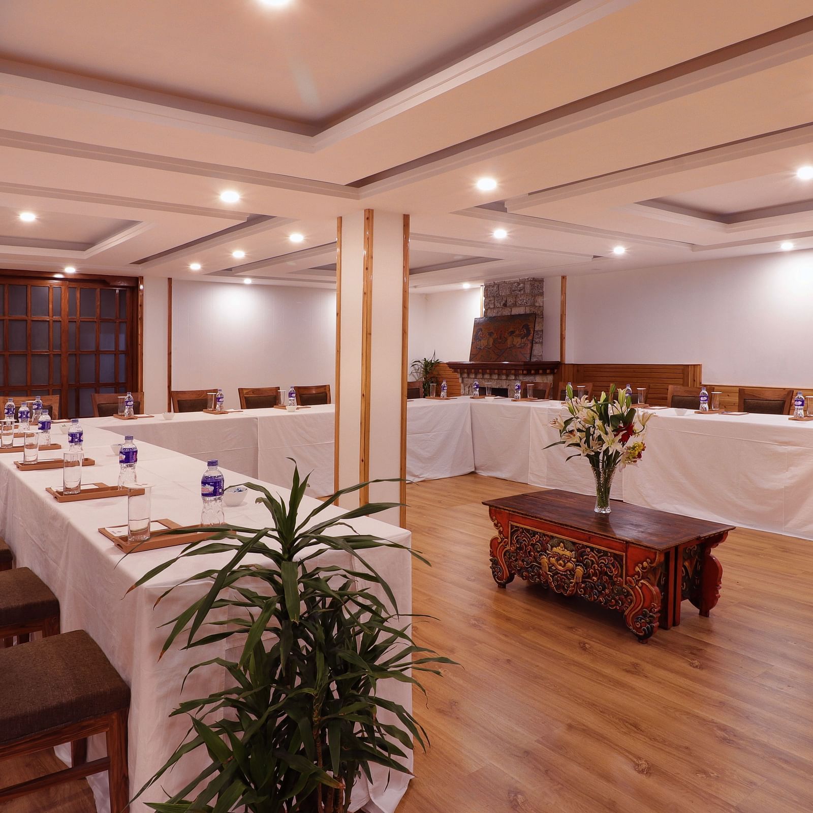 A view of the banquet hall at Denzong Shangrila Lords Inn Gangtok, featuring arranged elongated table and chairs with a carved wooden table and floral centrepiece in the centre.