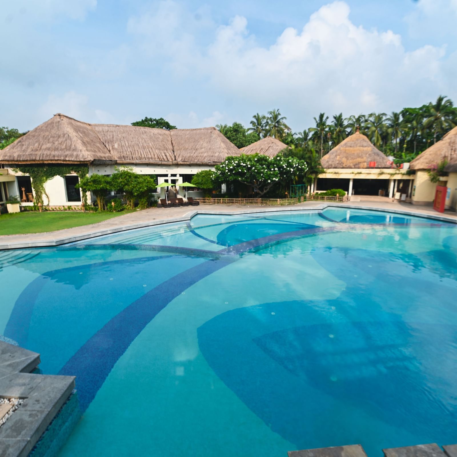 A daylight view of the common swimming pool at Vedic Village Spa Resort, featuring buildings with tatched roofs in the background.