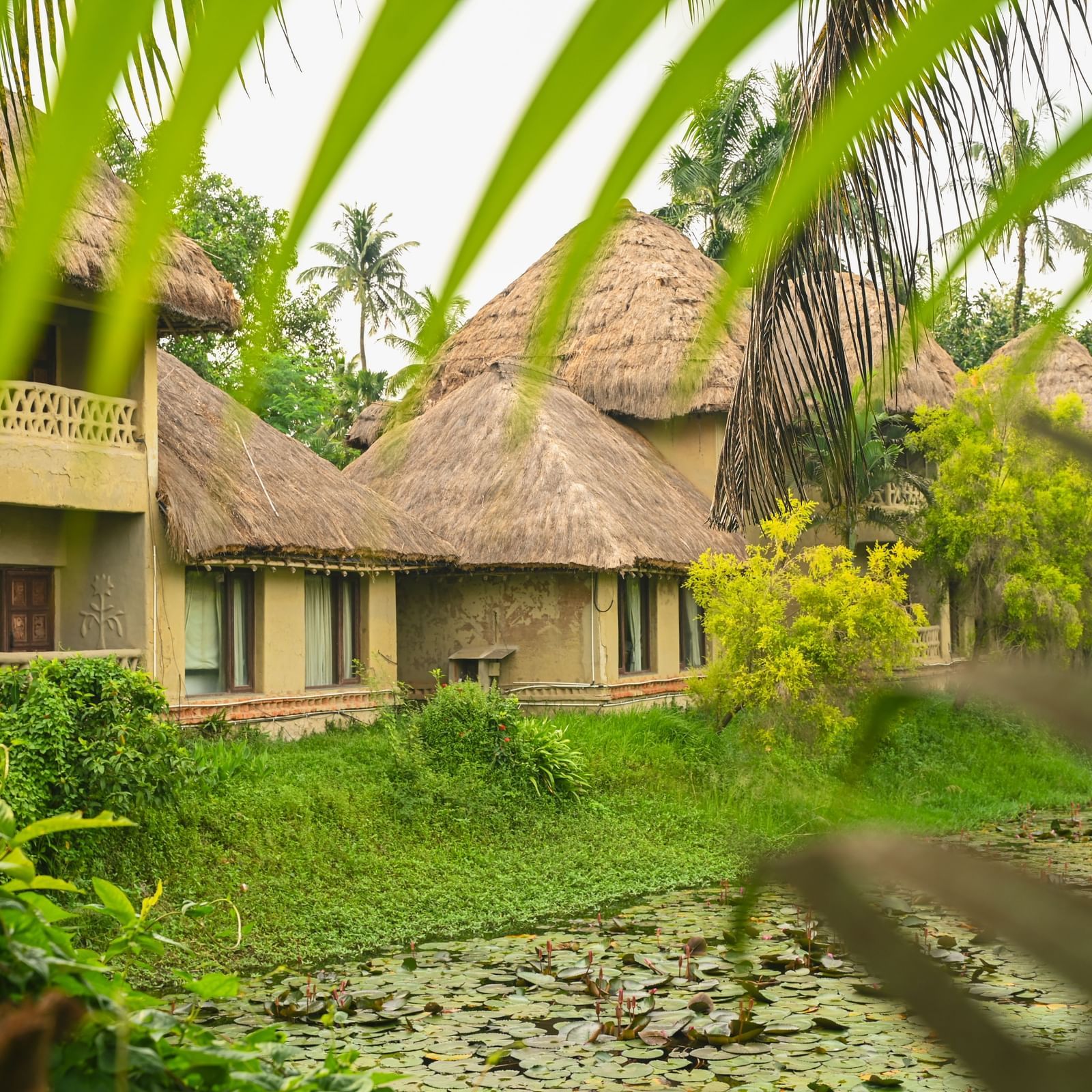 Exterior view of Earth Villa at Vedic Village Spa Resort showing mud-built structures with thatched roofs, set beside a calm water body and surrounded by dense greenery.