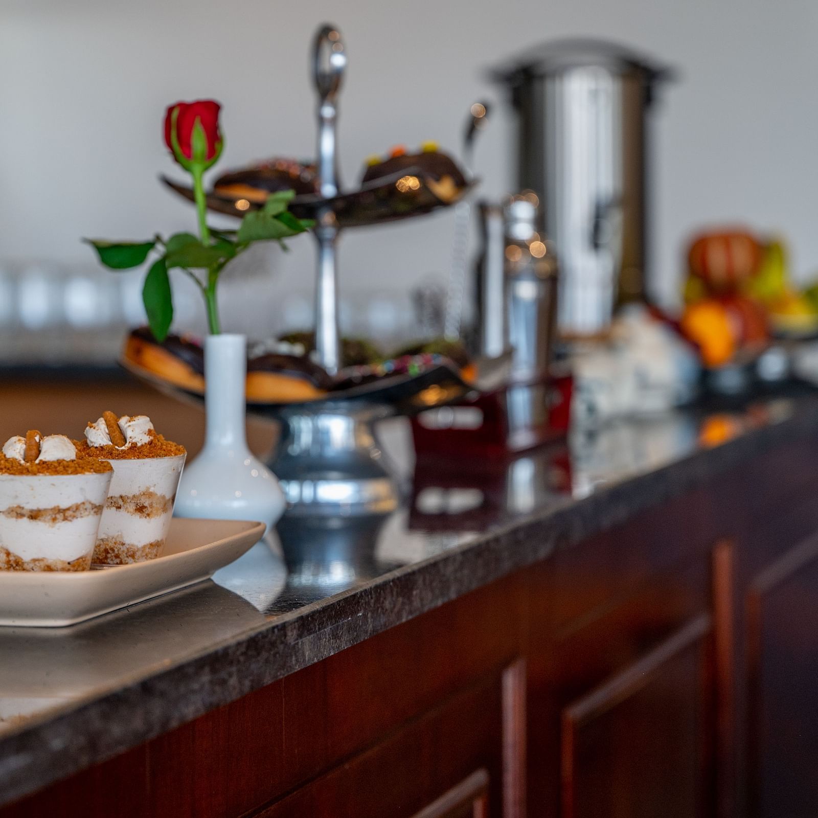 A dark wooden counter with a marble top holding various items, including a tray with two small desserts, a vase with a red rose and other bottles. - Vishranti Resort, Dehradun.
