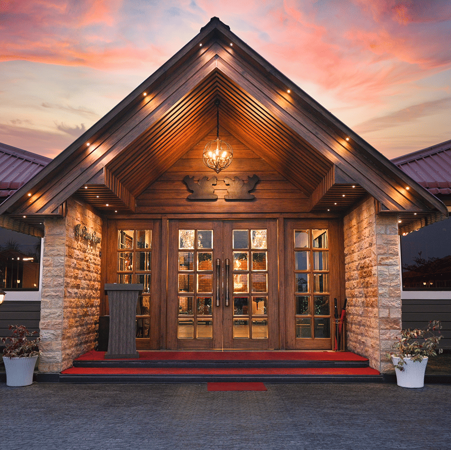 Piano Room entrance at dusk with a lit gabled wooden porch, glazed double doors, a red carpet and stone pillars at HM Hotel & Resort, Dibrugarh.
