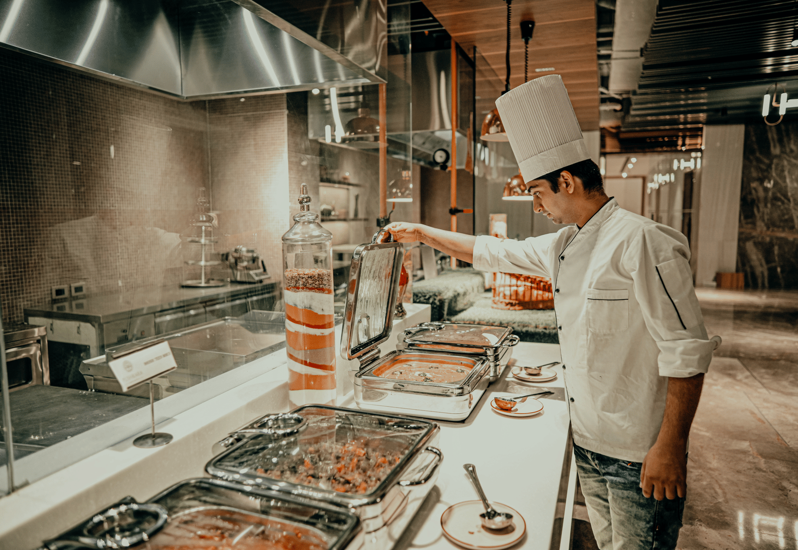 A chef in a white uniform prepares food behind a counter in a professional kitchen. There are different dishes in metal containers on the counter.