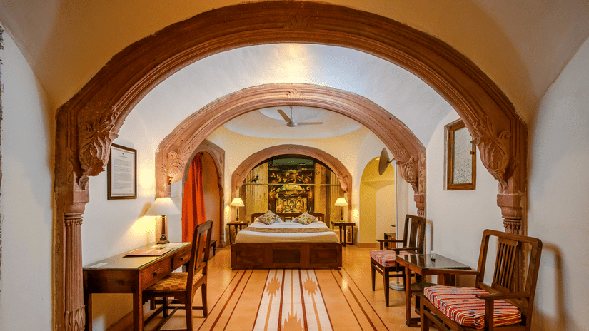 A hotel hallway with arched openings leading to a bedroom with a bed and other furnishings, as well as a desk and chairs along the hallway at Tijara Fort-Palace - 19th Century, Alwar.