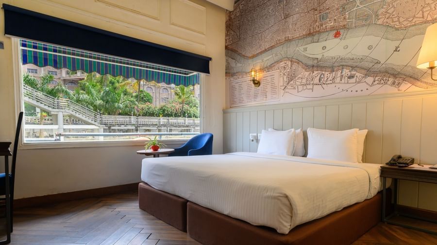 A modern hotel room featuring a large bed and a detailed map-like mural on the wall. A window with striped blinds overlooks lush greenery.