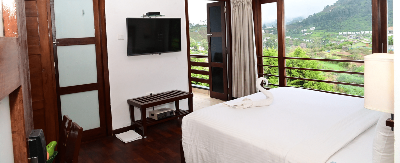 An image of a Rock Cabin Valley View with a king size bed, bed side table, lamps, flat screen TV, and an expansive window with curtains - GReaT trails Kodaikanal by GRT Hotels