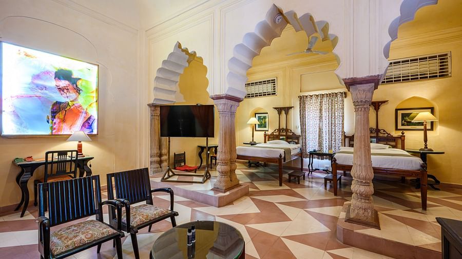A spacious room with two single beds, seating areas, a television, and arched architectural details at Tijara Fort-Palace - 19th Century, Alwar.