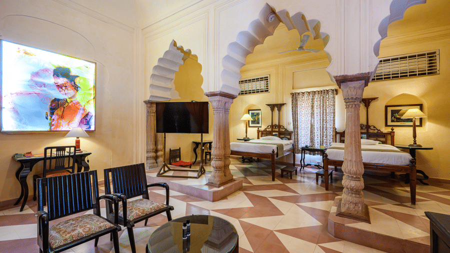 A spacious room with two single beds, seating areas, a television, and arched architectural details at Tijara Fort-Palace - 19th Century, Alwar.