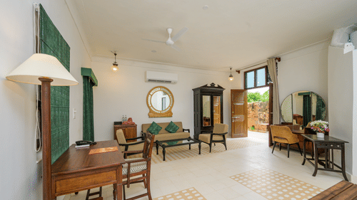 A spacious room with a dark wood desk, a seating area with a sofa, a round mirror, and an open doorway leading to an outdoor space, blending indoor and outdoor living with traditional decor - Tijara Fort-Palace - 19th Century, Alwar.