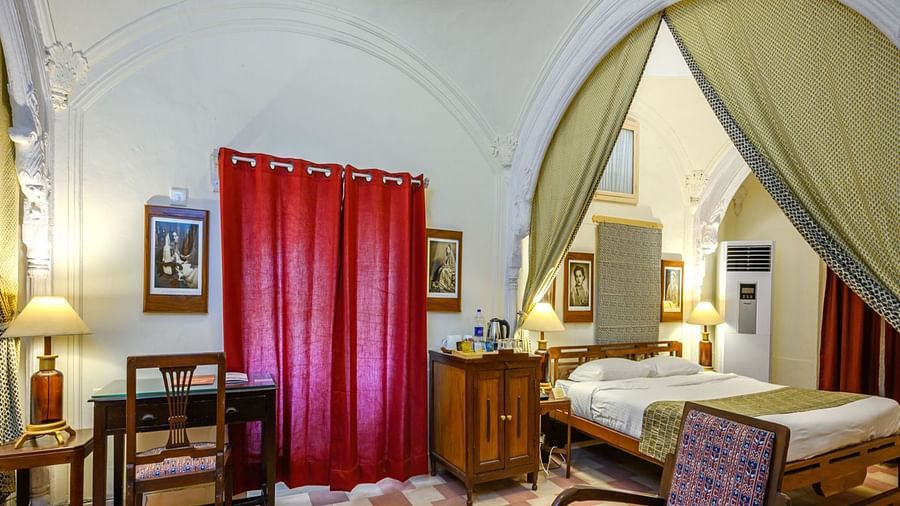 A bedroom with a bed enclosed by a decorative fabric canopy, a red curtain, a desk, and artwork on the walls at Tijara Fort-Palace - 19th Century, Alwar.