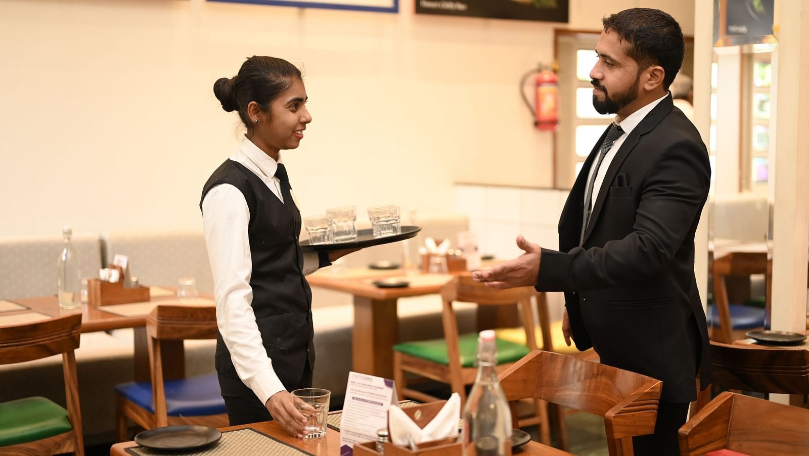 A waitress holding a tray of glasses while preparing a table in a restaurant - Kamats India