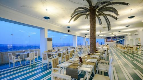 A dining area with multiple tables and chairs is visible inside Tijara Fort-Palace - 19th Century, Alwar, with large windows showing an outdoor view.