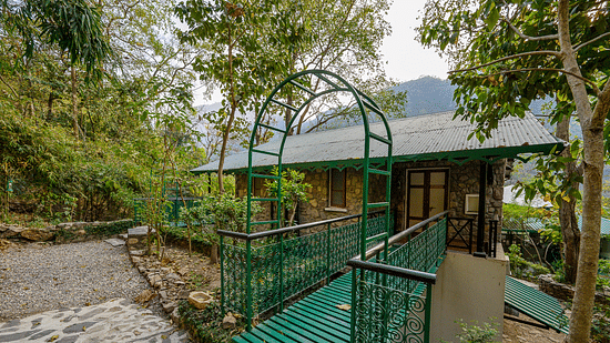 A cottage surrounded by lush greenery, featuring a green metal bridge-style entrance and cobbled pathways, nestled in a serene forest setting with distant mountain views.