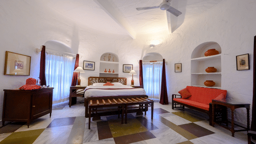 Hill Fort Kesroli 14th Century Alwar - interior view of the Mubarak Mahal room with a queen size bed, a ceiling fan and a red sofa with paintings on the wall