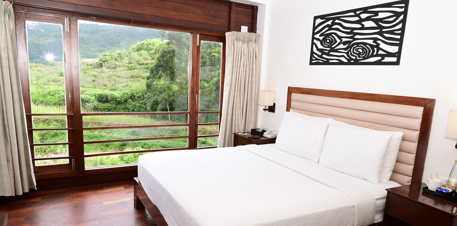 An image of a Rock Cabin Valley View with a king size bed, bed side table, lamps, wall paintings, and an expansive window with curtains - GReaT trails Kodaikanal by GRT Hotels