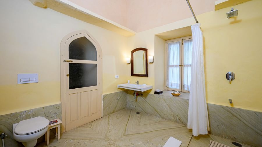 A bathroom with a sink, mirror, toilet, shower curtain, window, and a door with an arched top at Tijara Fort-Palace - 19th Century, Alwar.