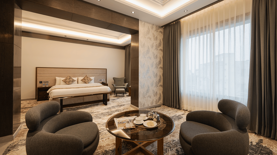 A spacious guest suite with a king-sized bed and two modern grey armchairs around a coffee table at Ananta Express, Katra.