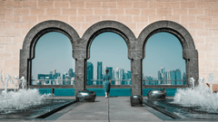 A woman walking through arches on a wall with doha's sky line in view