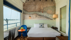 A hotel bedroom with a large bed, blue armchair, and a window with a view of the water and a lamp post. A vintage map mural is on the wall.