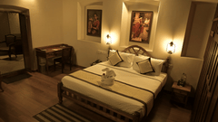 Heritage Suite at Uday Suites under warm lighting featuring a plush bed with bath towels made into an elephant on the bed runner and a work desk near the bed facing the wall while the wall above the headboard features a couple of paintings by Raja Ravi Var