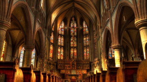 Inside of a church featuring benches, high ceilings and intricate architecture with stained glasses.