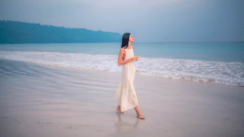 A woman in a white dress walks barefoot on a serene beach at sunset.