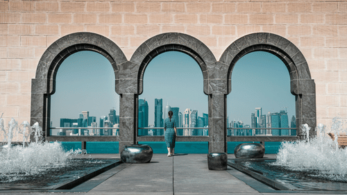 A person looking through 3 stone arches at a city skyline across water, with fountains in the foreground.