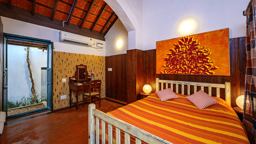 A cosy bedroom featuring a bed with striped orange and yellow linens, wood-panelled walls, an arched ceiling, and an open doorway leading to a small courtyard at Neemrana's Coconut Alley - 21st century, Tranquebar.