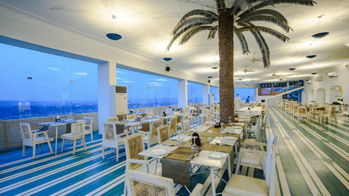 A dining area with multiple tables and chairs is visible inside Tijara Fort-Palace - 19th Century, Alwar, with large windows showing an outdoor view.