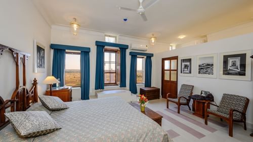 Room with bed, patterned pillows and bedding, furniture including chairs, tables, and a trunk, framed artwork, curtains, decorative ceiling lights, and patterned floor tiles - Tijara Fort-Palace - 19th Century, Alwar.