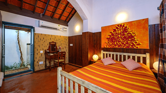 A cosy bedroom featuring a bed with striped orange and yellow linens, wood-panelled walls, an arched ceiling, and an open doorway leading to a small courtyard at Neemrana's Coconut Alley - 21st century, Tranquebar.