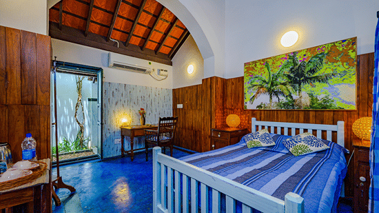 The King Room at Neemrana's Coconut Alley in Tranquebar, featuring a cosy bed with a painting on the wall above the headboard at a distance from the work desk next to the doorway