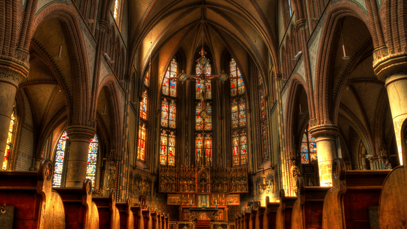Inside of a church featuring benches, high ceilings and intricate architecture with stained glasses.