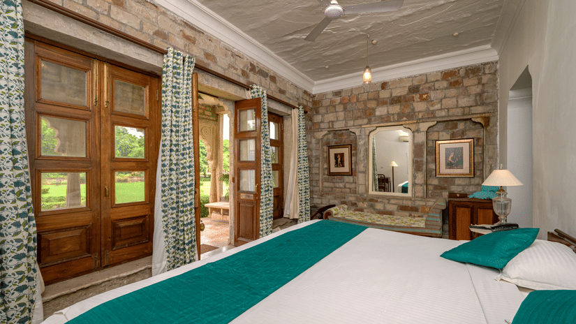 Spacious heritage bedroom featuring carved wooden doors, stone walls, patterned curtains, elegant furnishings, and a large bed overlooking lush gardens at Deo Bagh, Gwalior