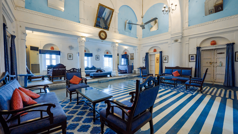Hill Fort Kesroli 14th Century Alwar - interior view of the Darbar Hall featuring a blue sofa set, white and blue painted walls, a ceiling fan and a chandelier