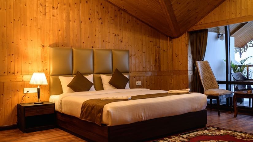A wooden bedroom featuring double sized bed at Istana Resort and Spa by Sagar, bedside table with a lamp, and a balcony attached where chairs are placed.