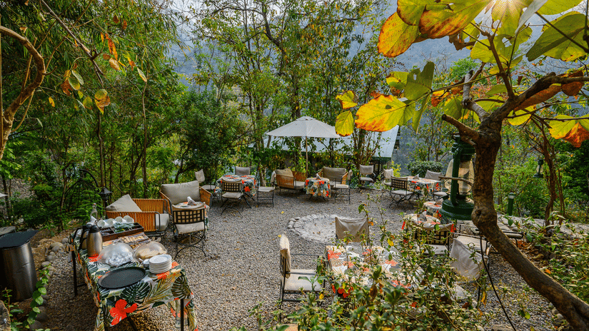 An outdoor dining area set amidst lush greenery, featuring wooden tables and a gazebo at Neemrana's Glasshouse on The Ganges.