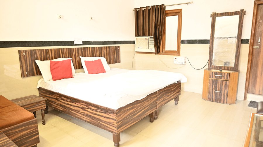A well-lit hotel room with a wooden bed frame, bedding, pillows, a wooden dressing table, an air conditioning unit, and a window with curtains - Hotel Goverdhan, Agra