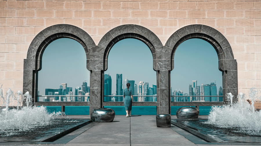 A person looking through 3 stone arches at a city skyline across water, with fountains in the foreground.