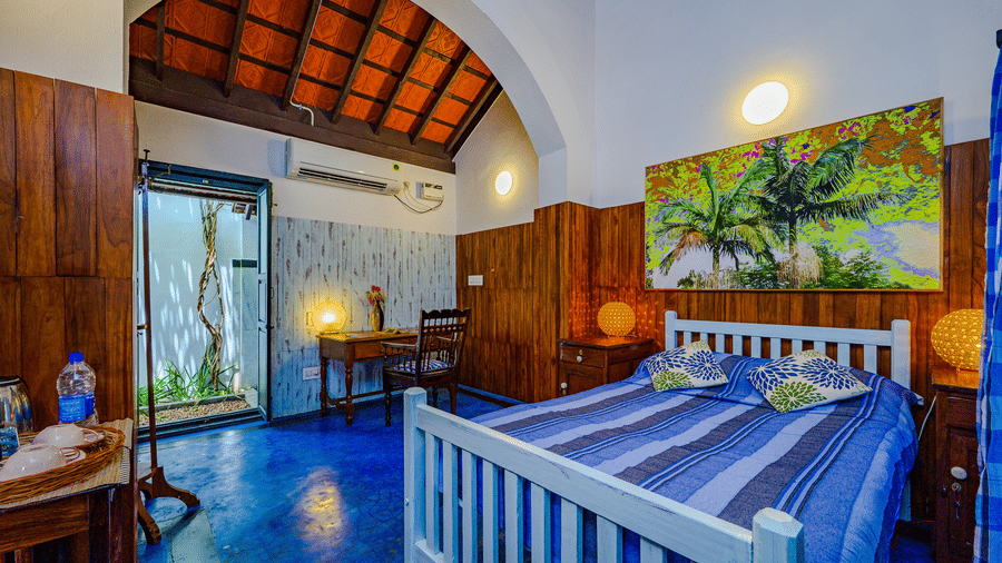 The King Room at Neemrana's Coconut Alley in Tranquebar, featuring a cosy bed with a painting on the wall above the headboard at a distance from the work desk next to the doorway