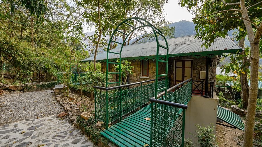 A cottage surrounded by lush greenery, featuring a green metal bridge-style entrance and cobbled pathways, nestled in a serene forest setting with distant mountain views.