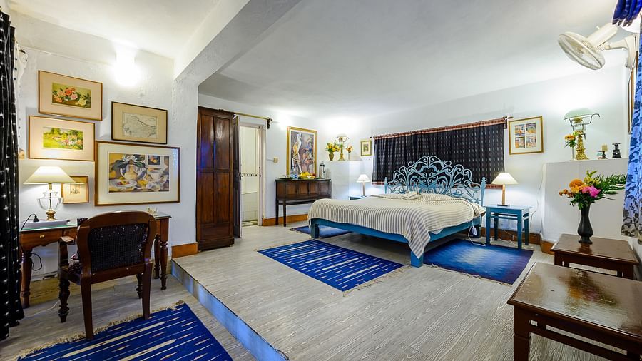 A grand room in a blue theme, featuring a work desk and wall paintings along with room amenities like an electric kettle, fruit basket, bedside lamp, etc. - Ramgarh Bungalows, Nainital.