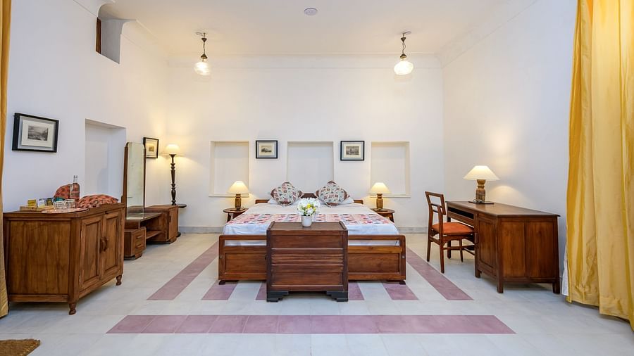 A hotel room with a bed featuring patterned bedding, wooden furniture including a dresser, a dressing table with a mirror, bedside tables with lamps, and a desk with a chair at Tijara Fort-Palace - 19th Century, Alwar.