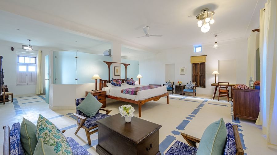Room with four-poster bed, patterned runner and pillows, furniture including chairs, tables, desk, and trunk, decorative ceiling lights, and patterned floor tiles - Tijara Fort-Palace - 19th Century, Alwar.