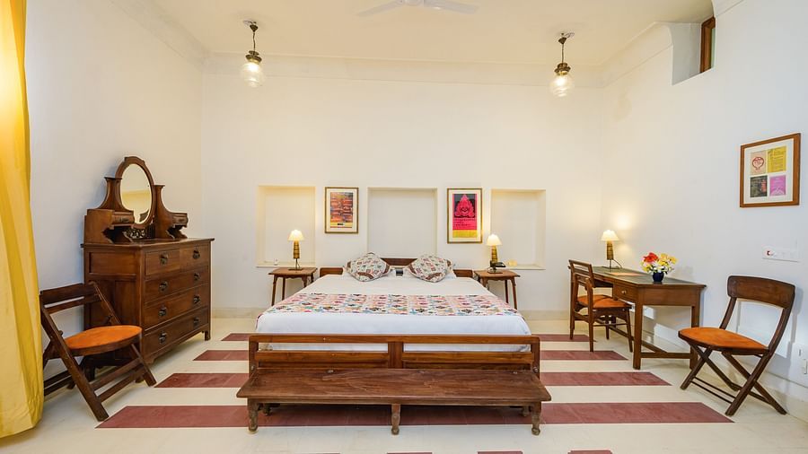 Room with bed, patterned pillows, furniture including desk, chairs, dresser, and bench, artwork, decorative ceiling lights, and patterned floor tiles - Tijara Fort-Palace - 19th Century, Alwar.