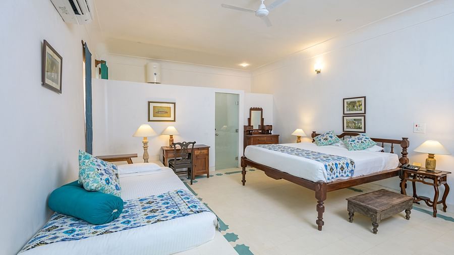 Room with two beds, patterned pillows and runners, furniture including desk, tables, and a trunk, artwork, wall-mounted air conditioner, and patterned floor tiles - Tijara Fort-Palace - 19th Century, Alwar.