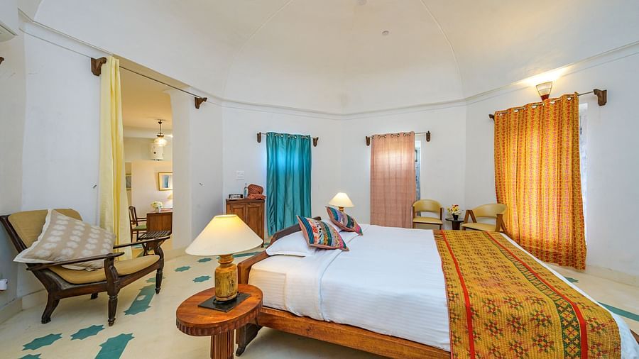 Room with bed, patterned pillows, curtains, windows, chairs, table, and lamp - Tijara Fort-Palace - 19th Century, Alwar.