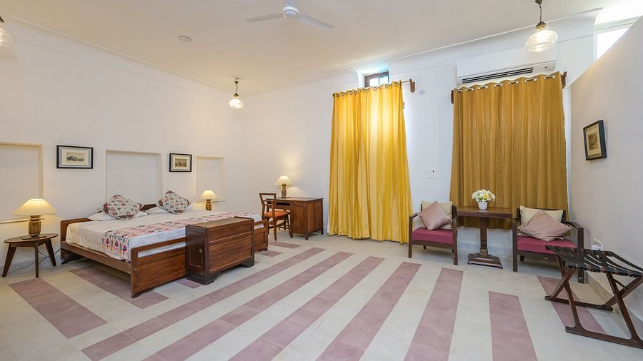 A spacious bedroom with a bed featuring patterned bedding, bedside tables with lamps, a desk with a chair, a seating area with chairs and a table, curtains on the windows, and framed pictures on the walls at Tijara Fort-Palace - 19th Century, Alwar.
