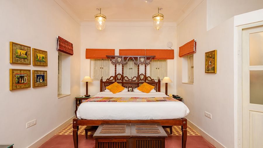 A bedroom with a wooden four-poster bed, orange accents, framed artwork on the walls, and a patterned rug, highlighting the room's traditional decor - Tijara Fort-Palace - 19th Century, Alwar.